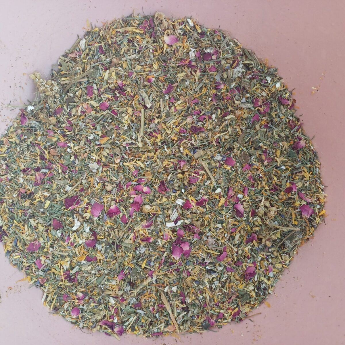 A high-angle close-up of a dry herbal blend in a pink bowl, featuring dried flower petals, green leaves, and various botanical ingredients.
