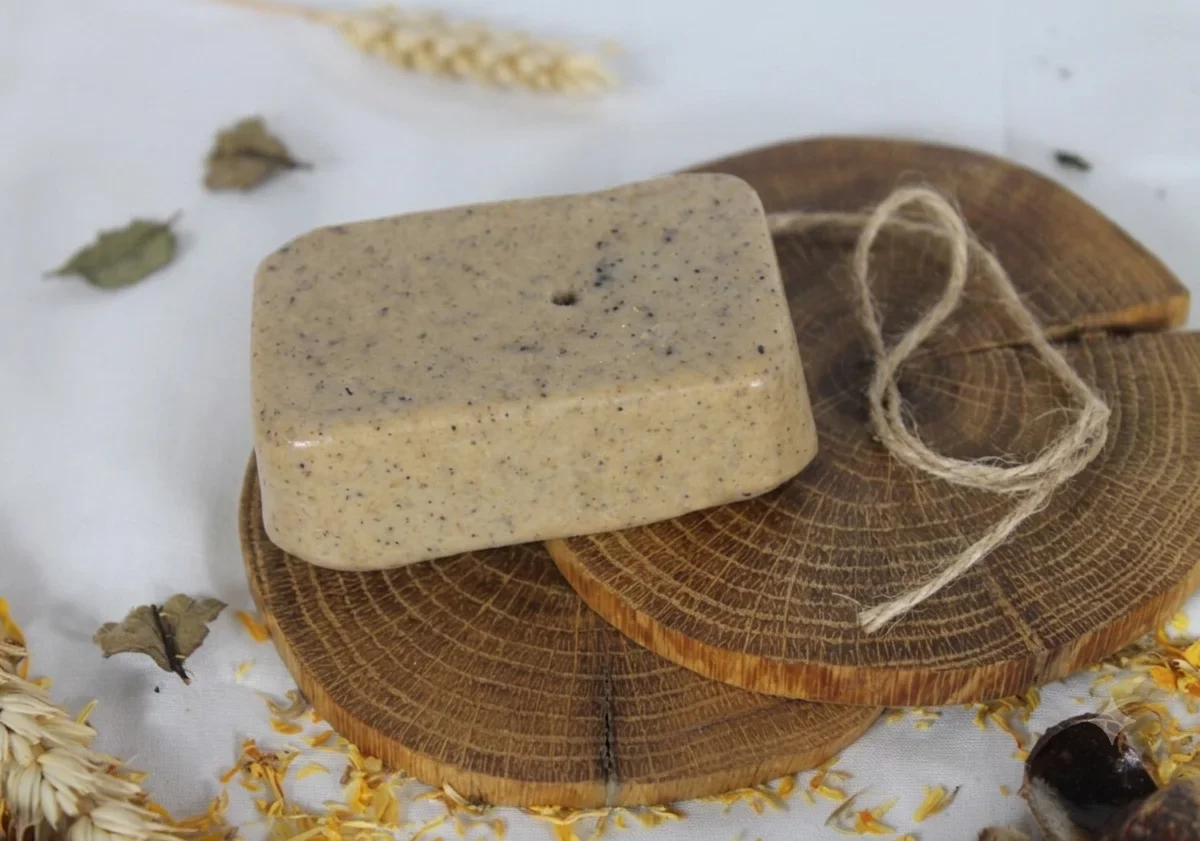 A handmade speckled exfoliating soap bar rests on wooden slices surrounded by dried wheat and flower petals. The minimalist display emphasizes the organic and botanical nature of the product.