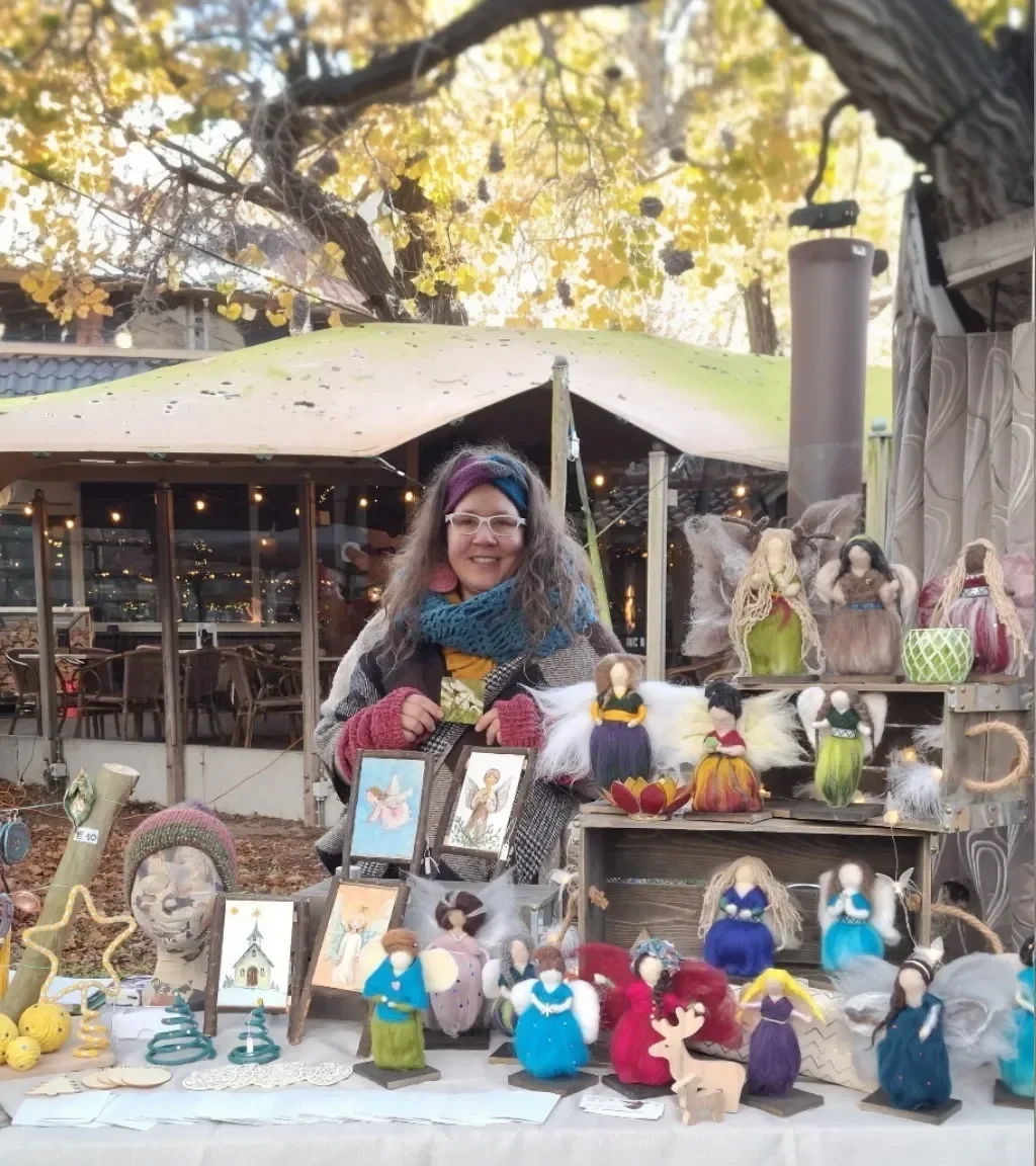 A woman stands behind a table displaying an array of handcrafted needle-felted fairy dolls and miniature forest-themed art at an outdoor autumn market.