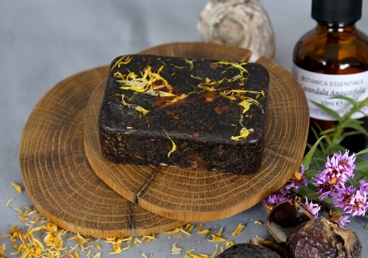 A block of handmade botanical soap topped with yellow flower petals rests on a wooden slice alongside a bottle of lavender essential oil. Dried botanicals and purple flowers decorate the surrounding scene.