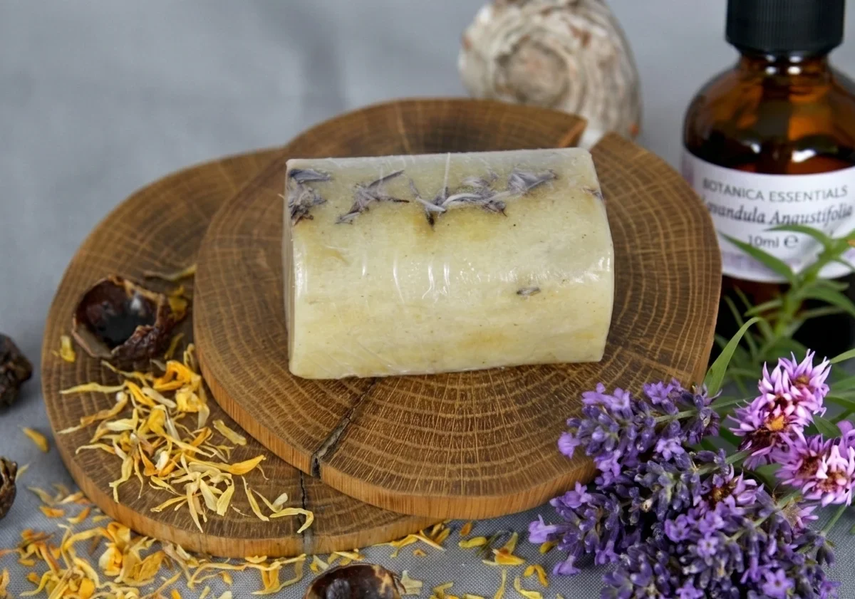 A handmade lavender soap bar wrapped in clear film sits on a rustic wooden slice, surrounded by dried petals and a bottle of essential oil. The scene emphasizes natural, handcrafted skincare beauty.