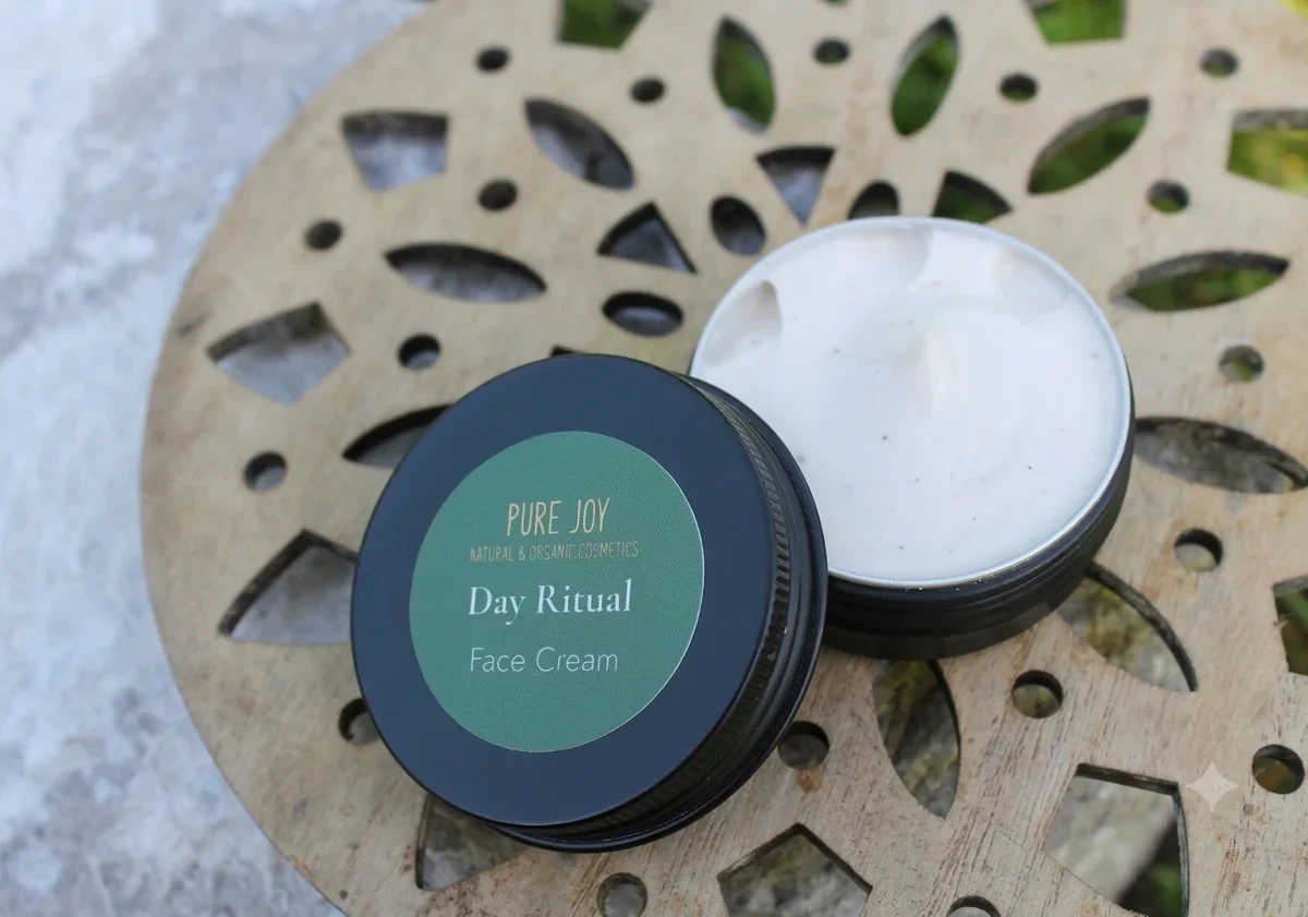 An open container of Pure Joy Day Ritual natural face cream sits on an intricate wooden mandala base outdoors. The cream is white with a smooth, rich texture, and the dark green label highlights the brand as natural and organic.