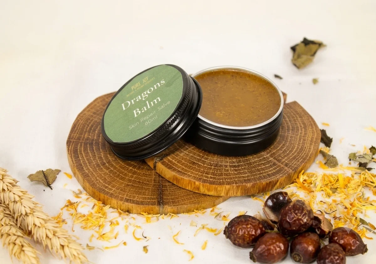 A jar of Pure Joy Dragons Balm skin repair salve resting on round wooden coasters, surrounded by rosehips and decorative wheat, representing natural handmade skincare.