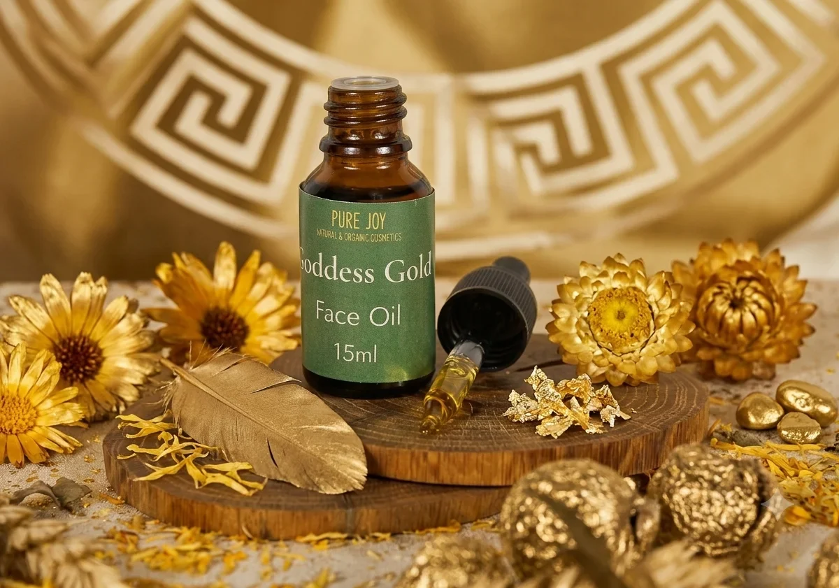 A bottle of Pure Joy Goddess Gold face oil sits on a wooden base with a golden feather and dried flowers. The aesthetic display highlights natural and organic skincare ingredients.