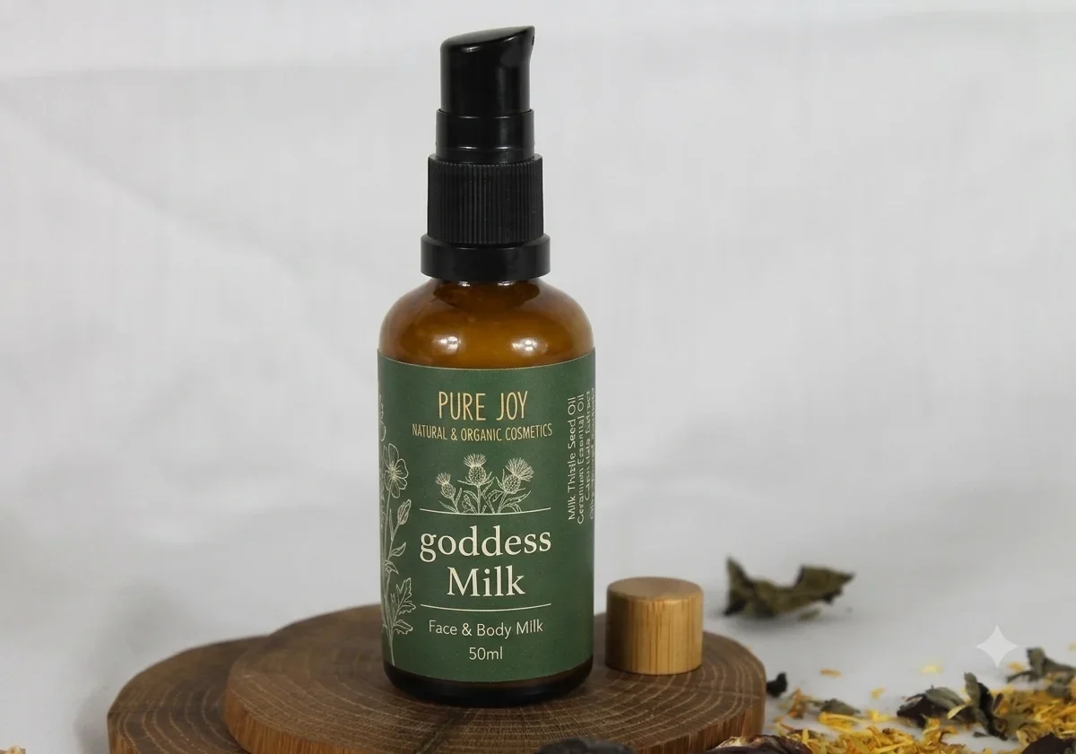A glass amber bottle of Pure Joy Goddess Milk face and body lotion sits on a circular wooden base, next to a small wooden cap. The scene is accented with scattered dried botanicals against a light background.