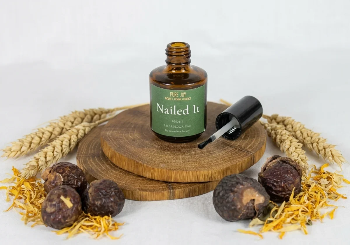 A small amber bottle of Pure Joy Nailed It organic nail treatment sits on two wooden disks surrounded by dried wheat, soap nuts, and botanical petals. This clean beauty product is presented in an earthy, sustainable display.