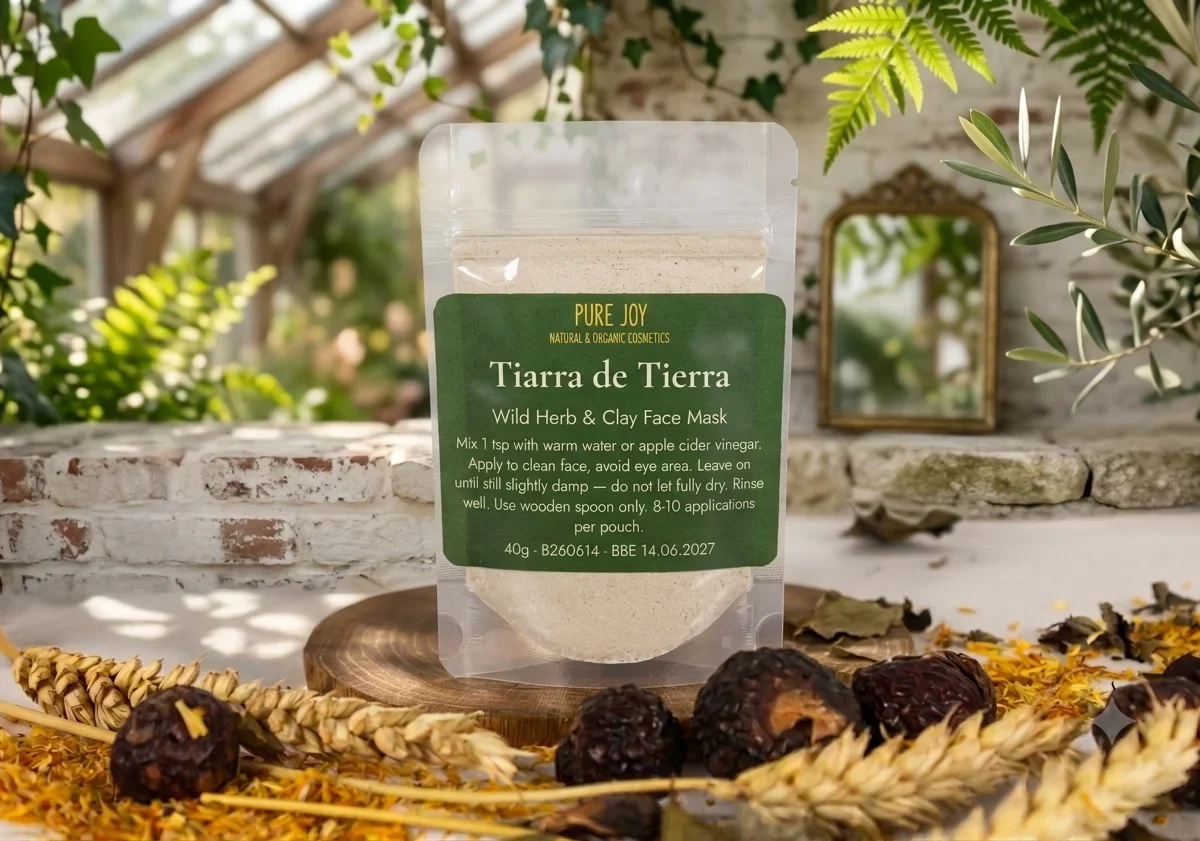 A pouch of Pure Joy Tiarra de Tierra wild herb and clay face mask sits on a wooden base surrounded by dried botanicals and wheat ears in a greenhouse setting.