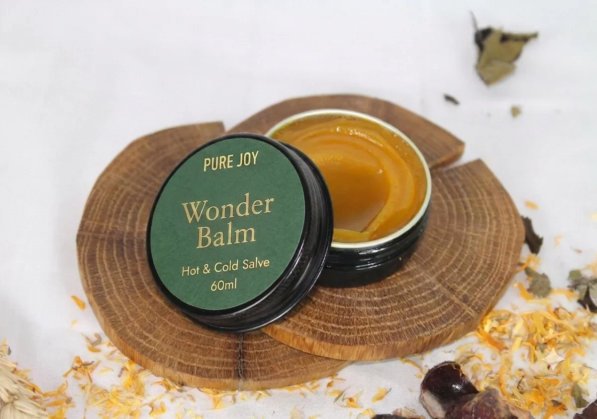 An open container of Pure Joy Wonder Balm, a natural hot and cold salve, sitting on a rustic wooden slice surrounded by botanical herbs.